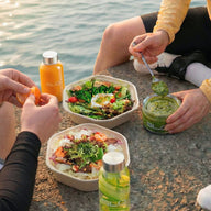 Two people enjoying a meal by the water, with salad bowls and bottles on a stone surface.