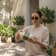 Person holding a green and white container outdoors with plants in the background